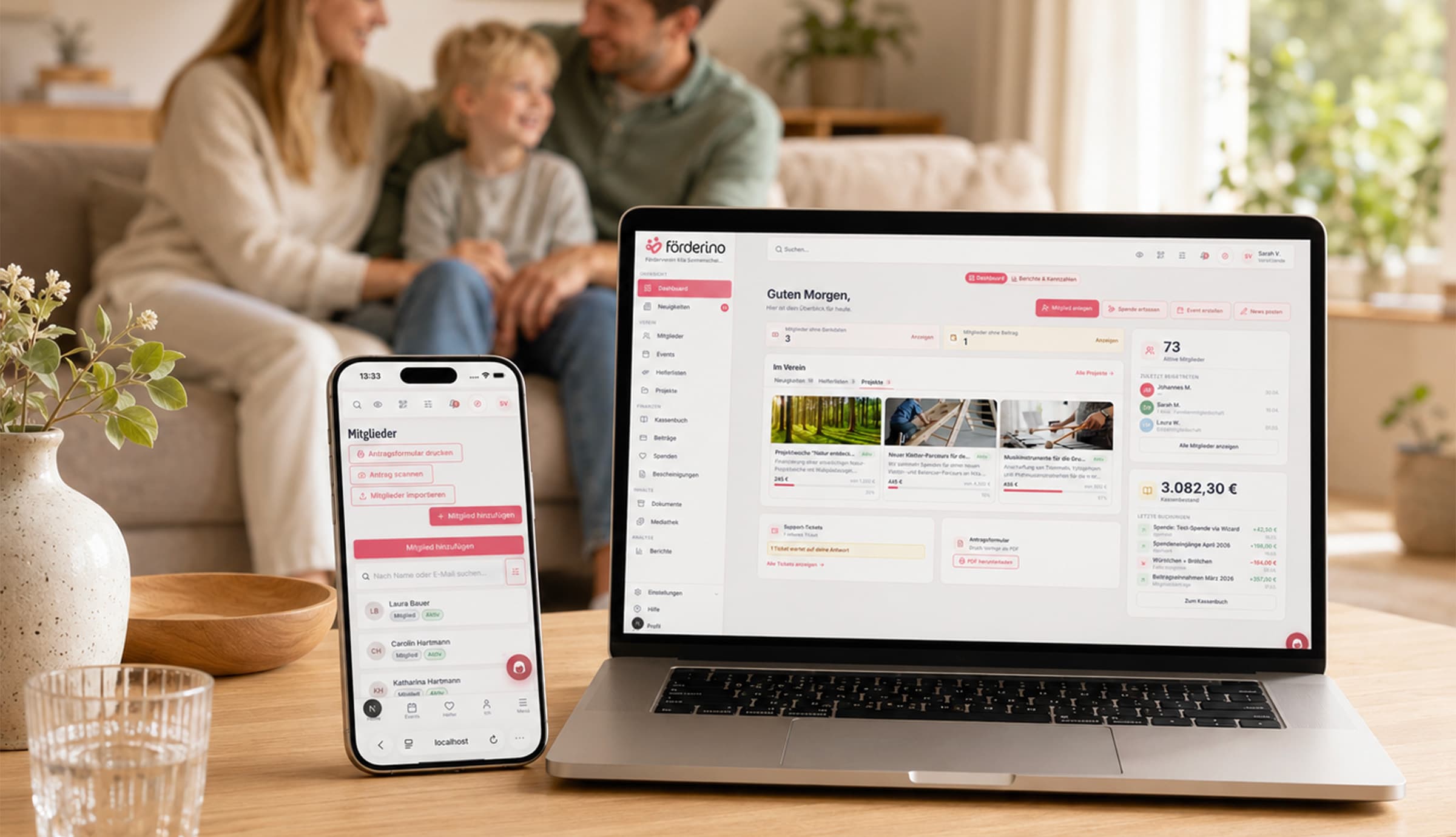 Auf einem Wohnzimmertisch stehen ein Laptop mit dem Förderino-Dashboard und ein Smartphone mit der Mitgliederliste, dahinter sitzen unscharf eine Frau, ein Kind und ein Mann auf dem Sofa.